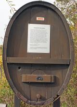 Frontansicht eines alten ovalen Weinfasses aus rotbraunem Holz. Im oberen Teil ist eine kleine Hinweistafel sowie ein Richtungspfeil angebracht.