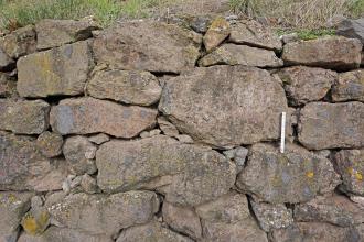 Teilansicht einer grauen bis rötlich grauen Stützmauer, errichtet aus unregelmäßig geformten Gesteinsbrocken. Als Füllmaterial zwischen den Fugen dienen kleinere Steinchen.