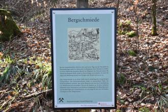 Blick auf eine kleine farbige Schautafel, aufgestellt am bergbauhistorischen Lehrpfad Birkenberg. Die Tafel zeigt eine Zeichnung sowie Erklärungen zum Thema Bergschmiede.