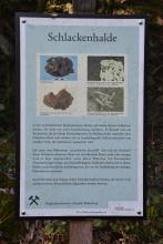 Blick auf eine farbige Schautafel, aufgestellt am bergbauhistorischen Lehrpfad Birkenberg. Die Tafel gibt Auskunft über Schlackenhalden und zeigt Fotos, etwa von verschlacktem Lehm oder Hammerschlag-Plättchen in Schmiedeschlacke.