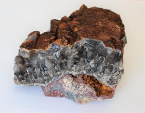 Großaufnahme von einem unregelmäig geformten Stück Rauchquarz. Das oben rotbraune Gestein zeigt vorne eine wellenförmige Bruchkante aus dunkelgrauen Kristallen. Am unteren Ende des Steins verlaufen mehrfarbige Marmorierungen.