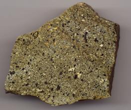 Nahaufnahme eines quadratischen Gesteinsbrockens mit oben dachartigen Schrägen und geschliffener Oberfläche. Das Gestein ist grünlich grau mit schwarzen, gelben und weißen Sprenkeln.
