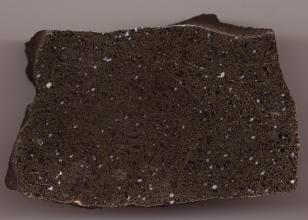 Großaufnahme eines rechteckigen Gesteinsbrockens mit gewellten Kanten und geschliffener Oberfläche. Das Gestein ist dunkelgrau mit kleinen schwarzen und weißen Sprenkeln.
