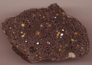 Nahaufnahme eines quadratischen Gesteinsbrockens mit schiefen Kanten und glatt geschliffener Oberfläche. Das Gestein ist violettgrau mit dunkelgrauen, gelben und weißen Einschlüssen.