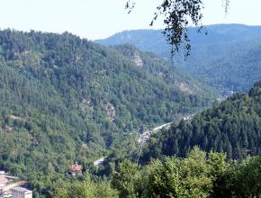 Blick von einem Berghang in ein enges, zwischen bewaldeten Bergen liegendes Tal. An den Berghängen, die nach links hin aufsteigen, ragen stellenweise Felsen hervor.
