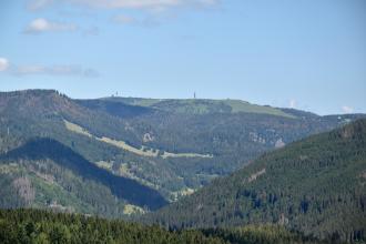 Blick von erhöhtem Standpunkt auf bewaldete Berge, die sich nach allen Seiten ausbreiten.  Im Hintergrund erhebt sich ein länglicher Berg mit kahler Kuppe, auf der ein Aussichtsturm zu erkennen ist.