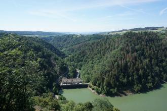 Blick aus großer Höhe auf mehrere bewaldete Berge, die links und rechts an den Ufern eines Sees aufsteigen. Der See hat grünliches Wasser und wird von einem Kraftwerk überspannt. Er breitet sich von unterhalb der Bildmitte nach rechts aus.