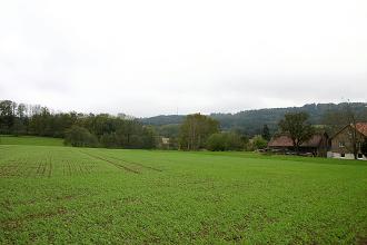 Blick auf bepflanzte Äcker, ein Gehöft rechts und Baumstreifen. Im Hintergrund bewaldete Höhenrücken.