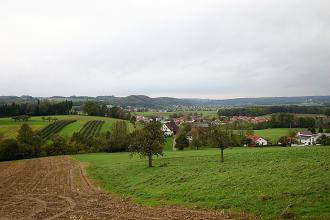 Blick über eine wellenartig geformte Acker- und Wiesenlandschaft mit Waldstreifen, einer Siedlung in der Mitte und bewaldeten Höhen im Hintergrund.