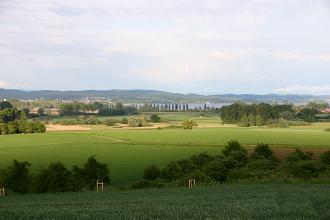 Blick von einem leicht erhöhten Standort auf eine flache Landschaft mit Wiesen, Bäumen und einer moorigen Senke. Im Hintergrund ein großer See, eine Stadt und bewaldete Höhen.