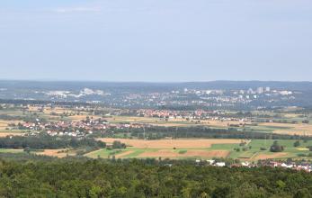 Aus beträchtlicher Höhe blickt man über flachhügeliges Gelände mit Äckern und vereinzelten Wiesen. Dazwischen verteilen sich Wälder und Siedlungen. Im Hintergrund liegt eine größere Stadt auf bewaldeten Hügelbergen.