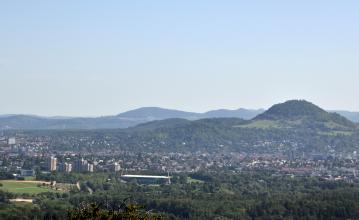 Vor bewaldeten Hügel und Bergen breitet sich eine größere Stadt aus. Der höchste Berg liegt rechts im Bild. Seine runde Kuppe ist bewaldet.
