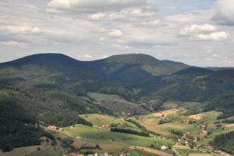 Blick in ein breites Kerbtal mit Wiesen, Äckern und einzelnen Häusern. An den Rändern des Tales erheben sich bewaldete Berge.