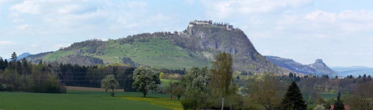 Das Panoramabild zeigt einen von links ansteigenden grünen Hügel, der rechts der Mitte in einen bewaldeten Gesteinskegel übergeht. Auf der flachen Kegelspitze trohnt eine Burgruine. Rechts hinten ist ein weiterer, spitzer Kegelberg erkennbar.