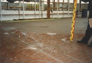 Das Foto zeigt aufgebrochenen und aufgewölbten Fliesenboden in einer alten Fabrikhalle. Rechts stellt eine Person eine Messlatte auf die Bruchkante des Bodens.