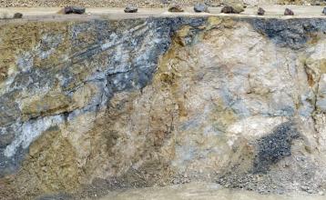 Blick auf eine 20 Meter hohe Abbauwand in einem Steinbruch. Das anstehende Gestein rechts im Bild ist hell, links davon befindet sich dunkelgraues und dunkelgelbes Gestein. Die verschiedenen Gesteine sind miteinander verwachsen.