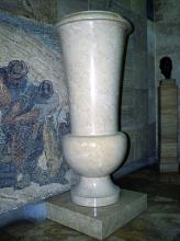 Das Bild zeigt eine sehr große, beigefarbene Vase. Die aus drei unterschiedlichen Teilen (Fuß, rundes Mittelstück und trichterförmige Öffnung) bestehende, marmorierte Vase steht auf einem niedrigen Podest.