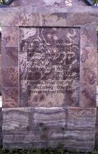 Blick auf den unteren Teil eines steinernen Denkmals mit eingravierten Namen. Das Gestein ist violettgrau und unten längs gestreift.