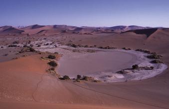 Das Bild zeigt eine afrikanische Wüste mit Dünen im Hintergrund und einem gewundenen, beinahe ausgetrockneten Fluss im Vordergrund. Der Fluss endet in einem runden, flachen Becken.