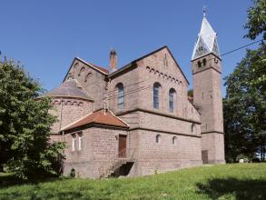 Blick auf eine in romanischem Stil erbaute Kirche mit Hauptschiff, hohem Turm rechts und kleineren Anbauten links. Die Kirche besteht aus rötlich grauem Gestein, die Dächer sind rot oder hellgrau.