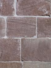 Nahaufnahme von rötlich grauem Mauerwerk mit schmalen hellen Fugen. Die Mauersteine sind rechteckig und weisen dünne Furchen auf.
