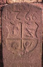 Blick auf einen hohen, aus rötlich grauem Stein gefertigten Grenzstein mit eingeprägtem Wappen und Jahreszahl.