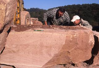 Das Foto zeigt einen rechteckigen rötlichen Steinblock mit unbehauenen, unregelmäßigen Kanten. Zwei Männer begutachten Oberfläche und Rückseite des Steinblocks.