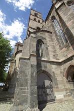 Ansicht einer Kirche in perspektivischer Verzerrung: Der Blick geht von unten nach oben. Zu sehen sind Turm (mittig oben) sowie Stützpfeiler und verschiedene Fenster. Das Mauerwerk der Kirche ist aus grauem bis rötlichem Gestein.
