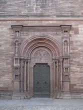 Eingangsportal einer Kirche mit Doppeltüre, schmalen Säulen und Verzierungen. Das verwendete Gestein ist von rötlich grauer Farbe.