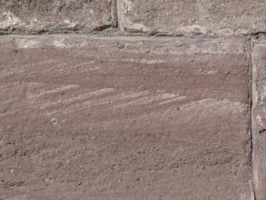 Nahaufnahme eines rötlich grauen Mauersteins mit teils waagrechter, teils schräger Schichtung sowie gröberen Poren unten. Oben und rechts sind weitere Mauersteine angefügt.