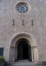 Frontansicht des Eingangs einer Kirche. Das dunkle Eingangstor wird von schmalen Säulen und runden Bögen umrahmt. Die Kirche ist aus rötlich grauen Steinen errichtet. Oben ist noch eine Fenster-Rosette zu sehen.