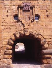 Blick auf einen tunnelartigen Mauerdurchgang aus rötlich braunen Steinen. Die Steine, die den Tunnelbogen bilden, sind klobig und abgerundet; die der Mauer rechteckig. Zwei runde Fenster sind oben um ein aufgesetztes Wappen in die Mauer eingelassen.