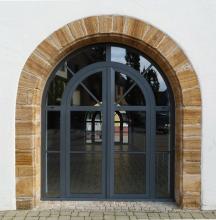 Blick auf ein modernes, aus Glas und Metall gefertigtes, oben abgerundetes Eingangsportal eines Gebäudes. Das Portal umgibt ein schmaler Kranz aus rötlichem bis grauem Gestein. Der sichtbare Rest der Fassade ist weiß.