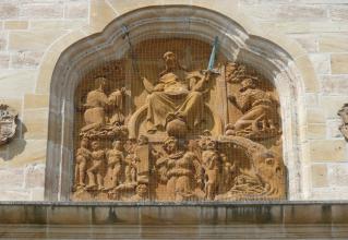 Das Bild zeigt eine aus rötlich braunem Gestein gefertigte Figurengruppe, untergebracht in einer fensterartigen Nische eines Gebäudes. Die Gruppe ist mit einem dünnen Gitter gesichert.