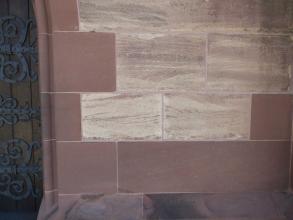 Mauer aus dunkelroten, homogenen Sandsteinblöcken und helleren, gebänderten Sandsteinblöcken. Am linken Bildrand ist ein Teil eines Eisentors zu sehen.