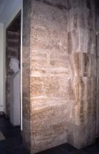 Blick auf bräunlich graue Gesteinsplatten, angebracht an Innenwänden eines Gebäudes. Rechts steht zudem ein Eckpfeiler aus gleichem Material. Links ist eine Nische mit einer hellen Steinfigur erkennbar.