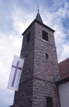 Blick von unten auf einen viereckigen Kirchturm mit spitzem Dach. Der Turm ist aus rötlichem Gestein gebaut. Auf der linken Seite des Turms ist eine weiße Fahne mit rotem Kreuz angebracht.