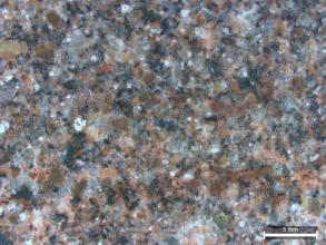 Detailaufnahme eines rötlich grauen Gesteins. Neben hell- und dunkelgrauen und rötlichen Kristallen finden sich auch ein paar helle Kristalle in dem Gestein. Rechts unten befindet sich ein Maßstab.