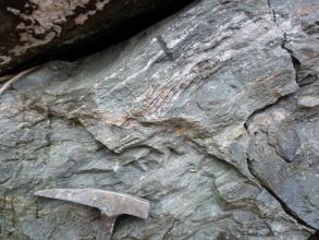 Detailaufnahme eines hell- bis mittelgrauen Gesteins, welches eine deutliche Einregelung der Kristalle in Bänder zeigt. Unten links befindet sich ein Hammerkopf als Maßstab.