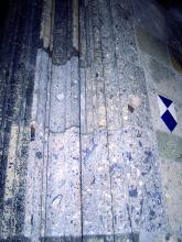 Das Bild zeigt Portalgewände und ein Stück einer Mauer in einer Kirche. Das Gestein ist bläulich grau (Gewände) bzw. hell- bis mittelgrünlich (Mauer) und weist viele Einschlüsse auf. Rechts auf der Mauer befindet sich noch ein blau-weißes Wappen.