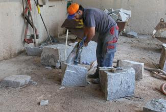Ein Mann in Arbeitskleidung und mit Bohr- oder Spaltmaschine zerkleinert graue Steinblöcke, die um ihn herum liegen. Im Hintergrund eine mit Steinen gefüllte Schubkarre.