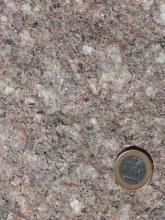 Nahaufnahme einer grobkörnigen, kristallinen Gesteinsoberfläche, Farbe rötlich grau mit größeren hellen Einschlüssen. Rechts unten liegt eine Euro-Münze.