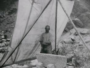 Sepiafarbenes Foto, das einen Mann beim Bearbeiten eines Gesteinsblockes zeigt. Links von dem Mann ist eine Leinwand aufgestellt, wohl zum Schutz vor der Sonne.