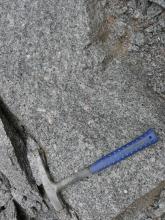 Nahaufnahme von grauem Gestein mit helleren Sprenkeln. Rechts oben ist die Oberfläche bröckelig, mit braunen Verfärbungen. Links unten ist eine schmale Spalte im Stein erkennbar. Daneben liegt ein Hammer.