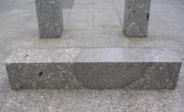 Nahaufnahme einer liegenden, vierkantigen Steinsäule. Farbe grau mit helleren Sprenkeln sowie dunklen Flecken. Der Stein liegt auf Steinplatten mit ähnlichem Muster. Im Hintergrund zwei aufrecht stehende Steinsäulen.