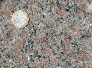 Nahaufnahme einer glänzenden Gesteinsoberfläche, Farbe grau mit rötlich braunen Kristallen. Links oben liegt eine Euro-Münze.