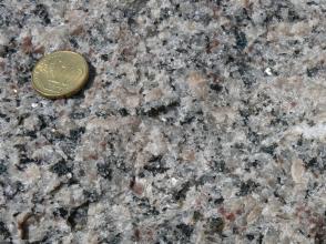Nahaufnahme einer kristallinen Gesteinsoberfläche, Farbe grau mit bräunlichen Stellen. Links oben liegt eine 10-Cent Münze. 