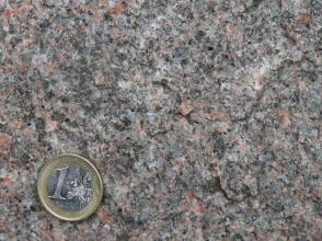Nahaufnahme einer kristallinen Gesteinsoberfläche, dunkelgrau mit rötlichen und vereinzelt weißen Einschlüssen. Links unten liegt eine Euro-Münze.