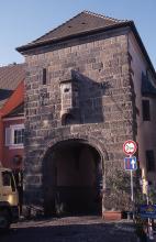 Das Foto zeigt ein altes gemauertes Stadttor als Turmbau mit Dach und bogenförmigem Durchlass sowie angebautem Erker. Die Mauersteine auf der Frontseite sind rötlich grau.