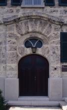 Blick auf den gemauerten Eingangsbereich eines Gebäudes mit Holztüre und ovalem Fenster. Das etwas vorstehende Portal aus gelblich grauem Gestein hat ein geschwungenes Dach und steht auf einem glatten grauen Sockel.